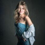 Portrait of a smiling blonde woman wearing a blue strapless top and a denim jacket draped over her shoulders, studio backdrop.