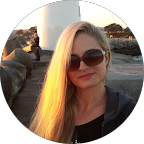 Portrait of a young woman with long blonde hair wearing sunglasses, posing outdoors near a rocky shoreline.